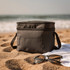 A grey double compartment cooler bag sits on sandy beach alongside sunglasses and a towel, with ocean waves in the background.