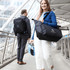 A black packable travel duffel bag held by a woman and a blue bag carried by a man, both on a city walkway.