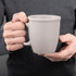 A ceramic mug in a light grey colour, held by a person with two hands. The mug has a smooth finish and a handle.