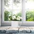 A white enamel metal cup filled with white flowers, placed on a rustic wooden surface near a sunny window.