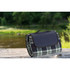 A picnic rug featuring a black and white plaid pattern, neatly rolled with a carrying handle, beside a river.