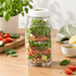 A glass salad jar filled with fresh vegetables, including lettuce, tomatoes, and cucumbers, featuring a logo.