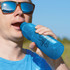 A drink bottle in a vibrant blue color, featuring a logo, is being held and used by a person wearing sunglasses.