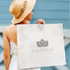 A beige garment bag with handles and a logo, held by a person wearing a straw hat and a white dress.