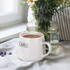 A white coffee mug with a handle, filled with coffee, beside a vase of flowers on a linen surface.