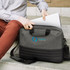 A grey and black laptop bag being opened, with a logo visible on the front. The person is seated on steps.
