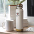 A drink bottle in white with a bamboo base, featuring a logo. A white mug and keyboard are in the background.