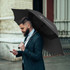 A black urban umbrella held by a man in a suit, standing outdoors while using a smartphone. The umbrella features a logo.