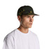 A camo cap in shades of green and brown worn by a model. The cap features a branded logo.
