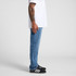 Straight blue jeans displayed on a standing model wearing a white t-shirt and black sneakers. The model has tattoos on their arm.