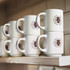 Stackable white coffee mugs displayed on a shelf, featuring a logo in brown.