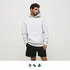 A man wearing a light grey hoodie and black shorts stands against a plain background. The hoodie has a logo.