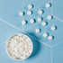A white bowl filled with round white mints, with several mints scattered around on a blue surface.