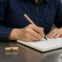 A cardboard pen in a natural finish, resting on a notebook beside a person writing with it.