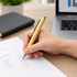 A bamboo pen in a hand, writing on a sheet of paper on a wooden desk, with a laptop and plant in the background.