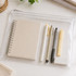 A clear pencil case containing a notebook, pens, a highlighter, and an eraser, all in neutral colours.
