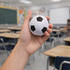 A hand holds a white and black soccer ball stress reliever in a classroom setting with desks and a chalkboard.