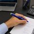 A blue recycled ABS pen is held above a notebook with notes, beside a laptop and calculator on a desk.