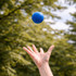 A blue Hi Bounce Ball is being tossed in the air against a blurred green background of trees.