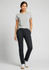 A woman stands in black chino pants and a grey t-shirt, smiling with one hand in her pocket. The image includes a logo.