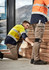 Two workers in high-visibility jackets and stretch cuffed pants, stacked wood planks in an industrial setting.