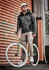 A man wearing cuffed beige stretch pants and a grey jacket stands beside a green bicycle against a textured orange backdrop.