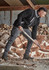 A model is wearing black work pants and a black fleece top while chopping wood in a rustic setting.