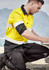 A man kneeling outdoors in a yellow and black long sleeve work shirt with reflective tape, engaged in construction work.