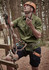 A man wearing a green short sleeve button-up shirt and blue shorts, equipped with climbing gear in a forest setting.