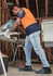 A man wearing an orange and navy short sleeve polo shirt works with a saw in a workshop setting.