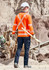 A worker in an orange long sleeve polo shirt with reflective stripes, holding a pickaxe, facing away from the camera.