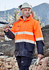 Hi-vis orange and navy waterproof jacket with reflective stripes, worn by a worker holding a tool on a construction site.