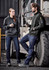 A man and woman wear black hoodies and blue denim work jeans, standing beside a large vehicle in an industrial setting.