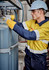 A worker in a yellow and navy spliced shirt adjusts equipment, wearing a hard hat and gloves in an industrial setting.