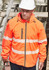 An orange packable rain jacket with reflective strips, worn by a person in a hard hat, standing near scaffolding.