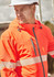 A bright orange unisex packable rain jacket with reflective stripes and a hood, worn by a man in a safety hat.