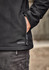 A black softshell jacket with a zippered pocket, featuring a logo and a hand tucked into the side.