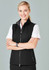 A woman in a black vest stands with her arms crossed, smiling against a neutral background. The vest features a zip.
