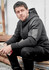 A black unisex zip front hoodie with multi-pockets, featuring a logo, worn by a seated man.