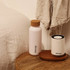 A drink bottle in pale pink with a cork lid, displayed on a wooden table alongside a white cylindrical object.