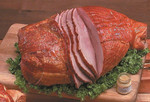 Cherry Smoked Sugar Cured Ham (Spiral Whole), sliced and served on a wooden cutting board with greens and a small jar of glaze.