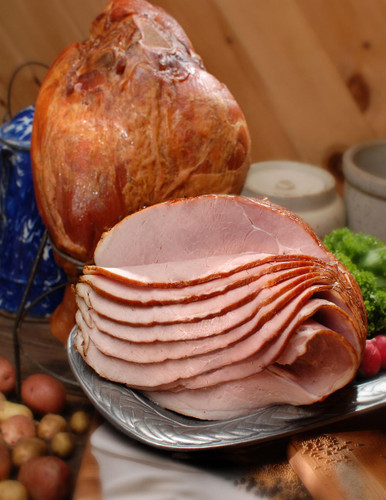 Cherry Smoked Sugar Cured Ham (Spiral Half), sliced ham with a pinkish interior on a silver platter, with whole ham in the background, surrounded by vegetables and food items.