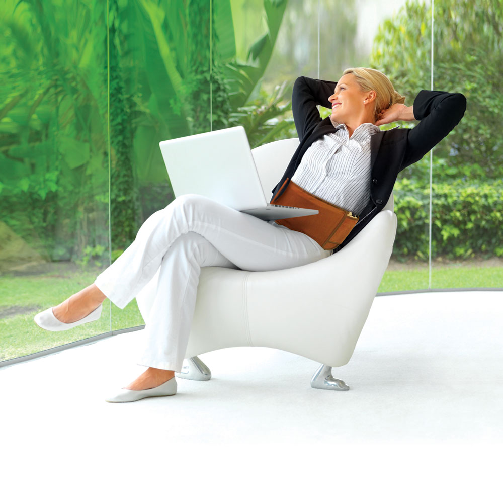 Woman sitting on a chair in a sunroom with vegetation outside.  She is smiling with a laptop on her lap and a Richway Biobelt around her waist with her hands behind her head.