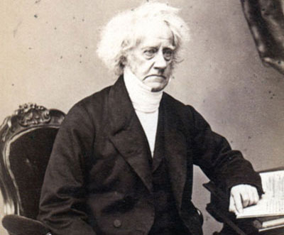 William Herschel sitting at a desk in front of a book with a pen in one hand