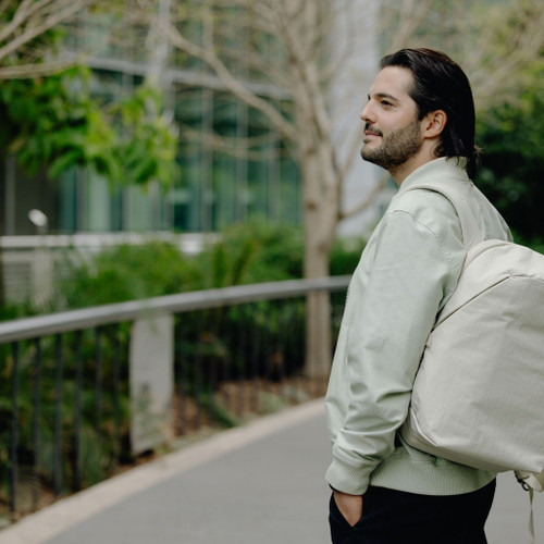 A man in a light sage green hoodie and black pants stands on an outdoor pathway, carrying a moon color backpack over his shoulder, with trees and a modern glass building in the background. | Peak Design official