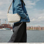 A person wearing a blue denim jacket and dark wide-leg pants carries a moon color 6L City Crescent bag with a thin strap, standing by a waterfront with a city skyline in the background. | Peak Design official