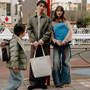 Three people standing on a city street beside a red pole, with a moon color tote bag hanging from the man's hand. The scene includes a woman in a beige sweater and lapis color backpack, a man in an olive jacket and brown pants, and a child in a green puffer jacket walking past. | Peak Design official