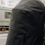 A close-up of a black backpack with a zippered pocket and a small 'piku design' label, shown outdoors with a blurred train in the background. | Peak Design official