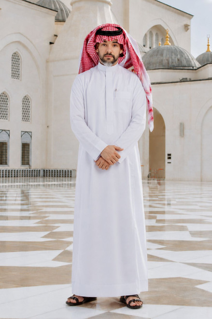 Man in traditional white thobe with red keffiyeh standing in front of a mosque.