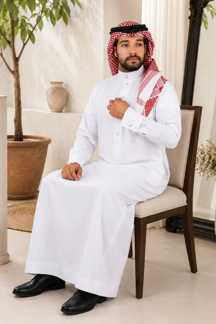 Man in traditional white thobe with red scarf sitting on a chair indoors.
